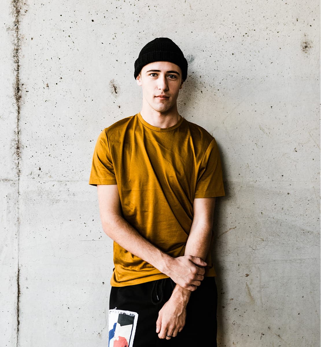 Молодой мужчина в шапке и футболке с коротким рукавом. Он опирается на бетонную стену.