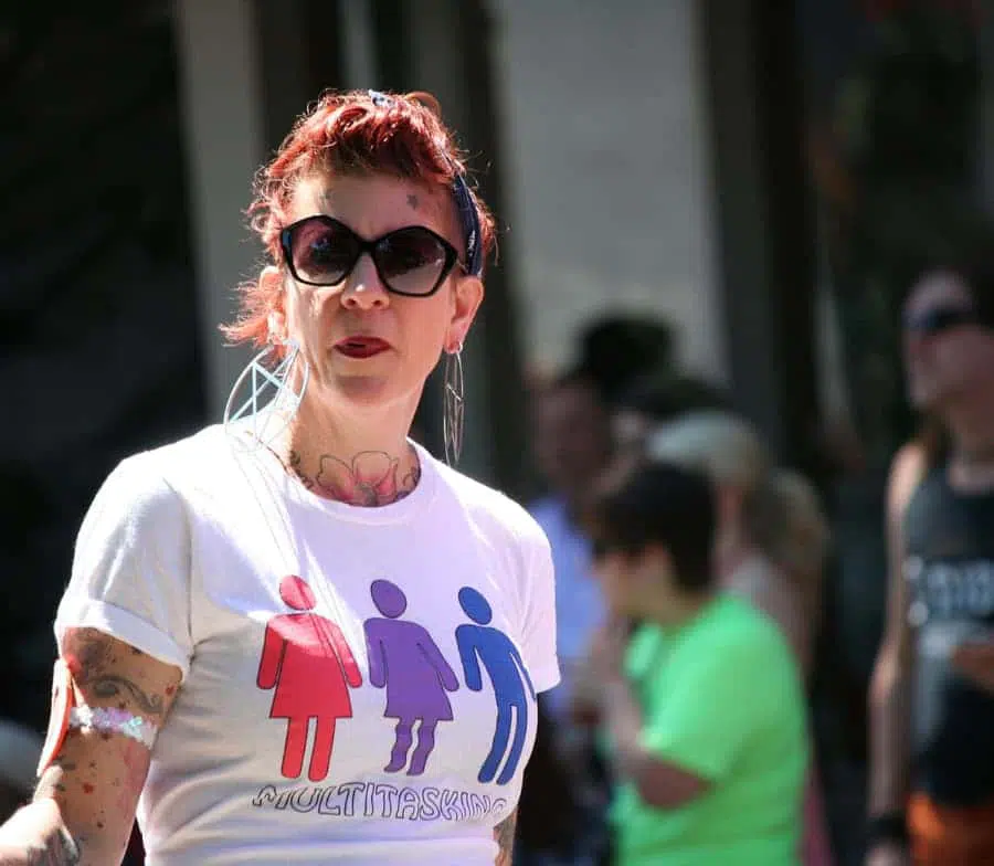 Frau mit langem zusammengebundenem Haar, Sonnenbrille und Tattoos auf den Oberarmen und auf dem Hals steht auf einem öffentlichen Platz.