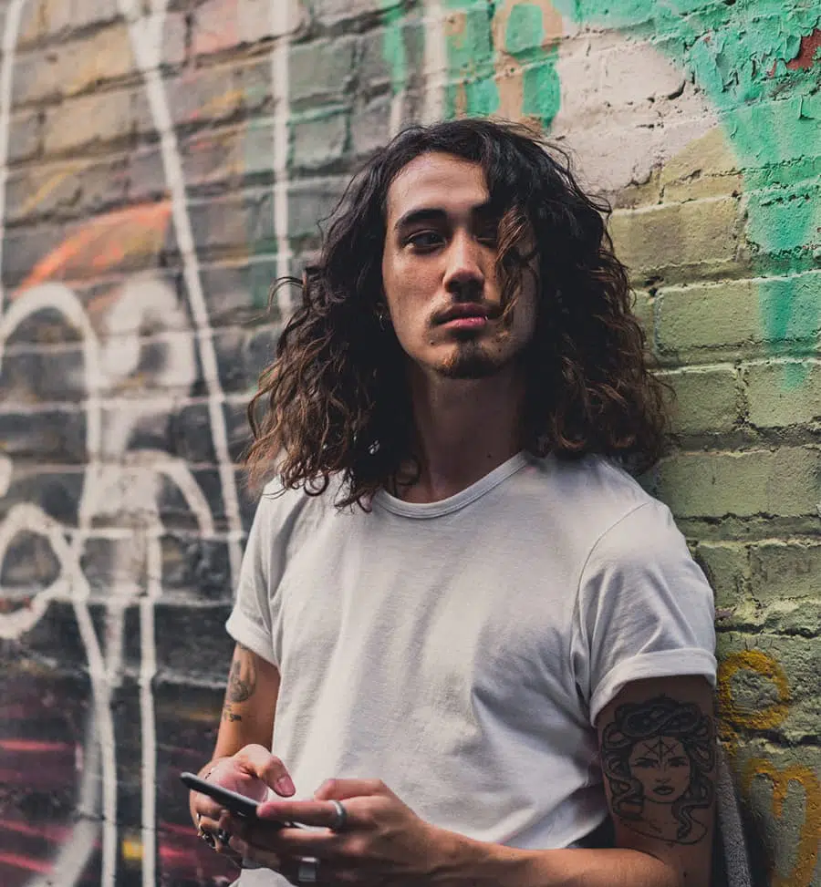 Junger Mann mit Kinnbart und schulterlangem Haar, ist angelehnt an eine Backstein-Wand mit Graffiti und hält ein Mobilfunkgerät in der Hand.